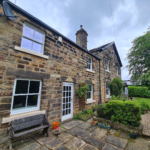 Old yellow stone house in Leeds with six new white sliding sash windows and a White timber door with sections. The house has a garden bench, hanging basket and a planter with hedging plants.