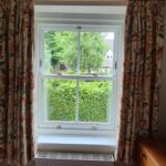 Brand new timber sliding sash window with antique brass fittings by Patchett Joinery. The window sits between two orange-patterned curtains.